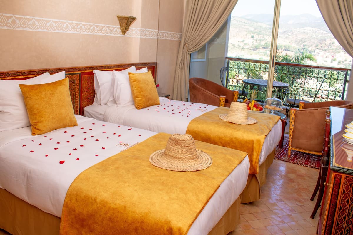 Habitación de hotel con dos camas, un balcón con mesa y sillas, y vistas al paisaje.