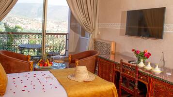 Habitación de hotel con cama, una mesa de comedor con un florero y televisión montada en la pared, con vista a la ciudad a través de la ventana.
