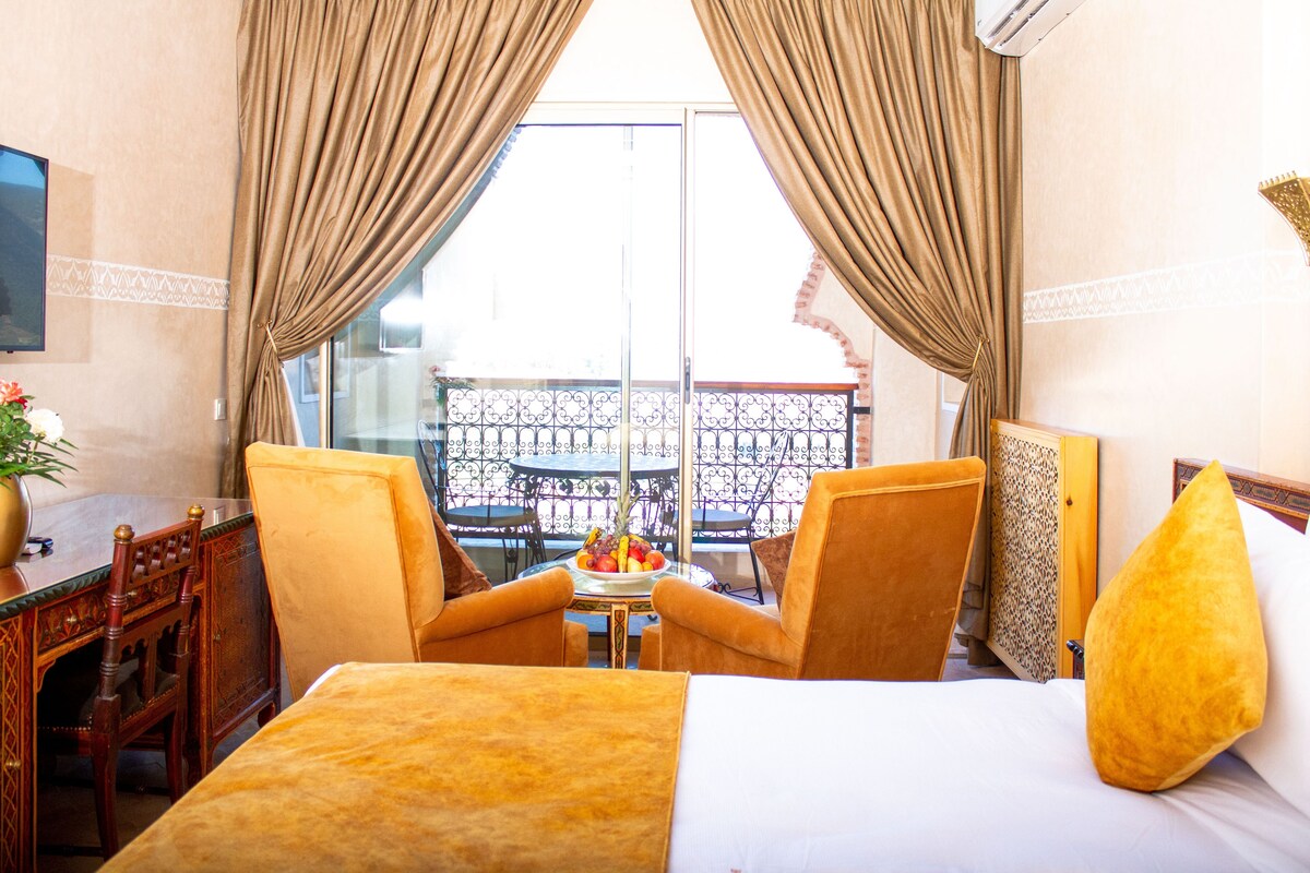 Ein Hotelzimmer mit einem Bett, zwei Stühlen, einem kleinen Tisch mit Obstschale und einem Balkon mit Geländer.