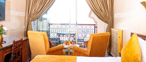 Une chambre d’hôtel comprenant un lit, deux chaises, une petite table avec un panier de fruits et un balcon doté d’une rambarde.