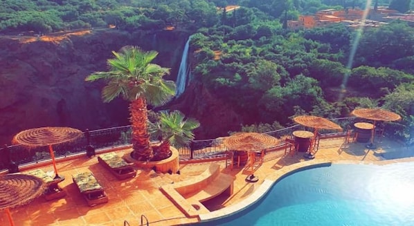 A poolside area with lounge chairs, umbrellas, and a waterfall in the background. - Borj Des Cascades & Spa (Ait Taguella)