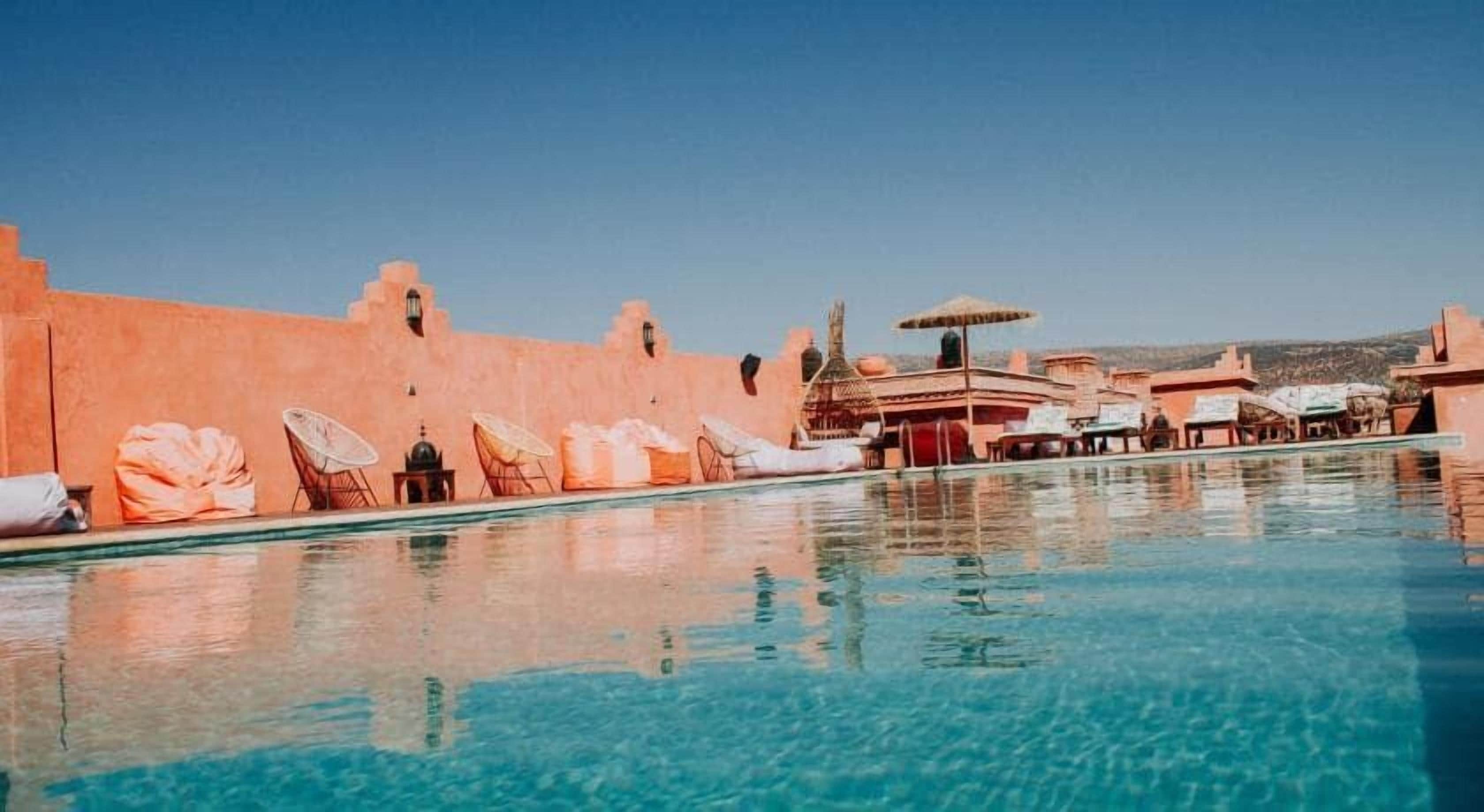 Una piscina con tumbonas y sombrillas, rodeada por un edificio de fachada rosa.