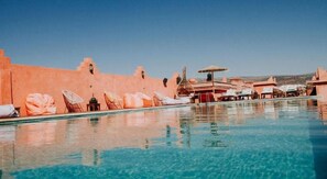 Ein Swimmingpool mit Liegestühlen und Sonnenschirmen, umgeben von einem Gebäude mit einer rosafarbenen Fassade.