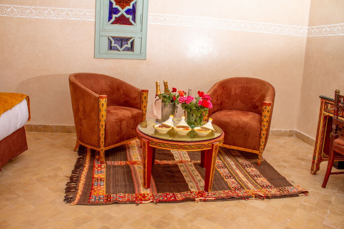 A room with two brown armchairs, a small round table with a vase of flowers, and a bed with a white cover.