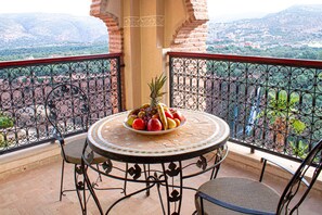 Een balkon met een ronde tafel en stoelen, een schaal fruit en uitzicht op een bergachtig landschap.