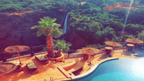 Un área junto a la piscina con sillones de descanso, sombrillas y una cascada al fondo.