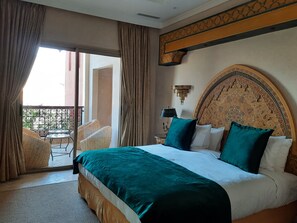 Habitación de hotel con una cama grande, un balcón con mobiliario de mimbre y un cabecero decorativo.