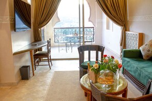 Ein Zimmer mit Balkon, einer Couch, einem Esstisch mit Stühlen, einem an der Wand befestigten Fernseher und einer Mülltonne.