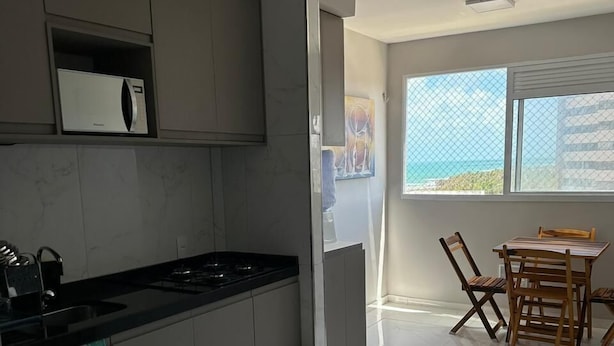 Eine moderne Küche mit Einbauherd, grauen Schränken und Fliesenboden. Ein Essbereich mit Tisch und Stühlen sowie ein Fenster mit Meerblick.