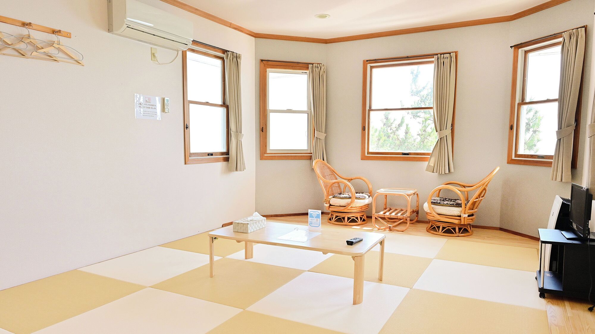 A room with a checkered floor, a table, two wicker chairs, a TV stand, and windows with curtains.