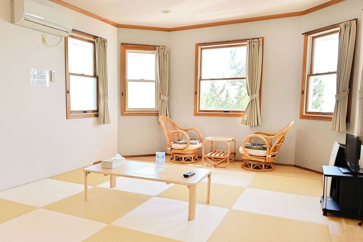 Ein Zimmer mit Schachbrettmuster-Boden, einem Tisch, zwei Korbstühlen, einer TV-Kommode und Fenstern mit Vorhängen.