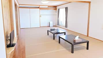 Quarto com piso de tatame, portas deslizantes, televisão, duas mesas e janela com cortinas.