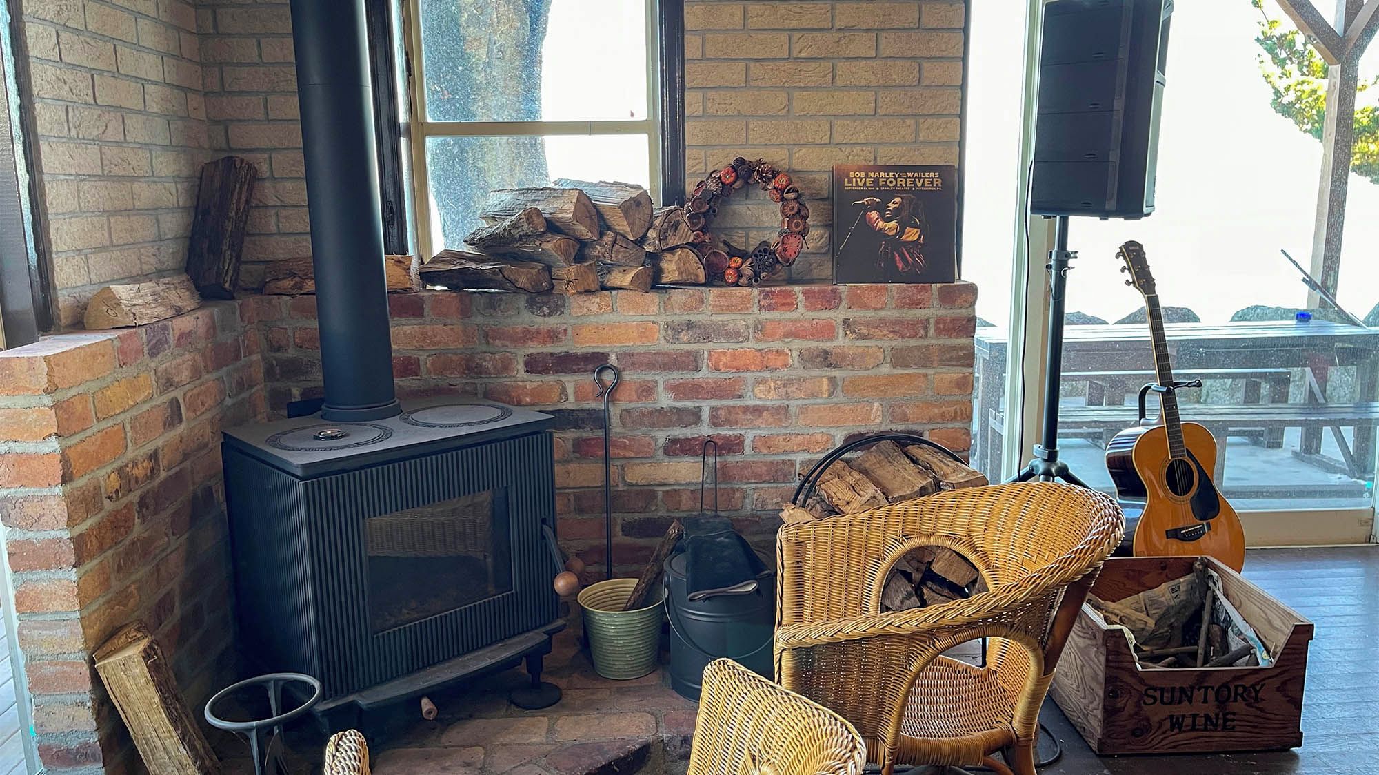 Una habitación acogedora con chimenea de ladrillo, mobiliario de mimbre, una guitarra y un cartel que dice 'SUNTORY WINE'.