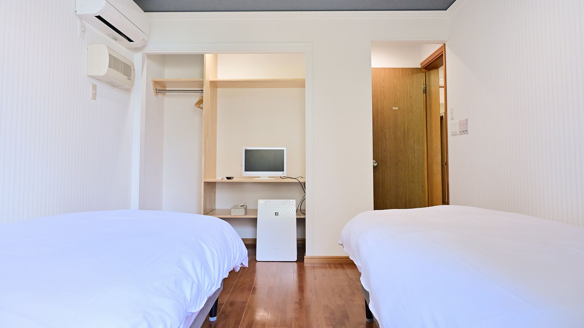 A hotel room with two beds, a desk with a computer, a wardrobe, and an air conditioning unit.