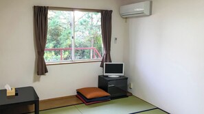 Een kamer met een raam, een televisie op een standaard, een klein tafeltje met zakdoekjes en een futon met kussens.