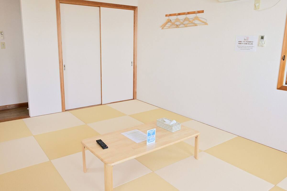 Ein Zimmer mit Tatami-Boden, einem Holztisch mit Fernbedienung und Taschentuchbox, einer Schiebetür und einem Wandheizkörper.