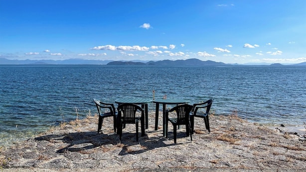 A table and chairs by a lake with mountains in the background. - B B Dylan (Otsu)