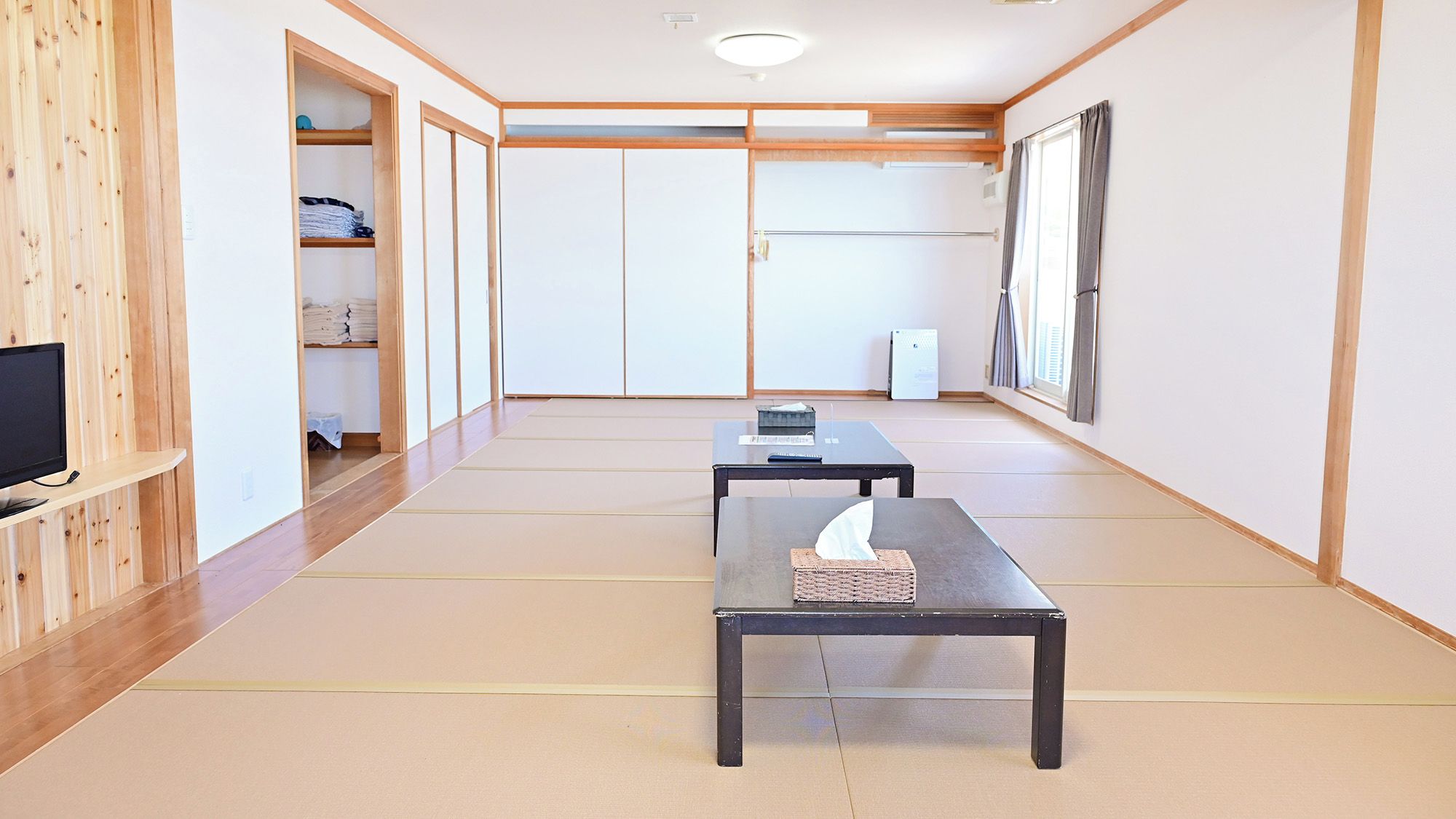 Una habitación con suelo de tatami, un televisor sobre una estantería, dos mesas, un armario y una ventana con cortinas.
