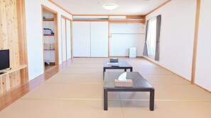 Une pièce avec un sol recouvert de tatamis, un téléviseur posé sur une étagère, deux tables, une armoire et une fenêtre avec des rideaux.