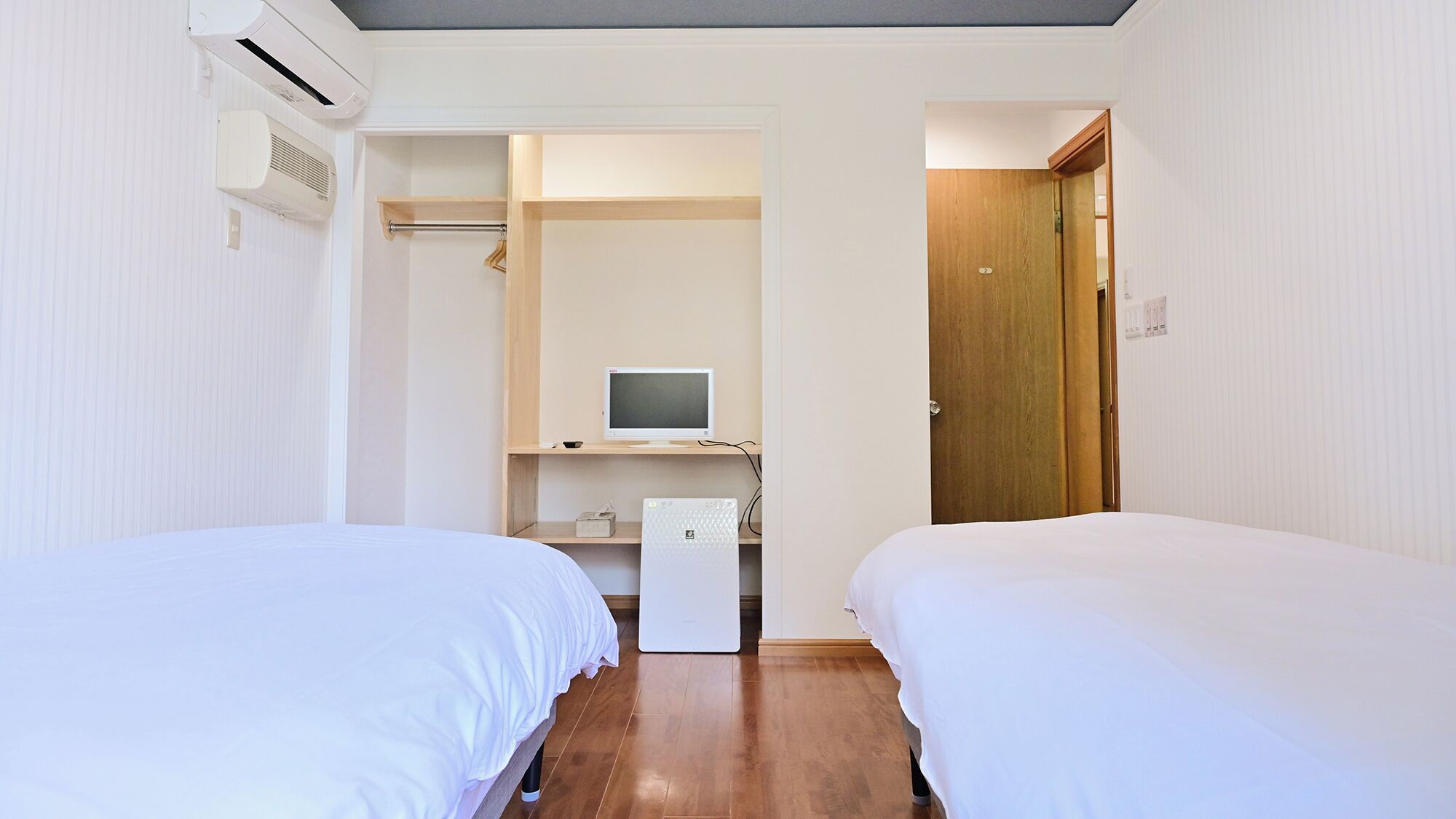 A hotel room with two beds, a desk with a computer, a wardrobe, and an air conditioning unit.
