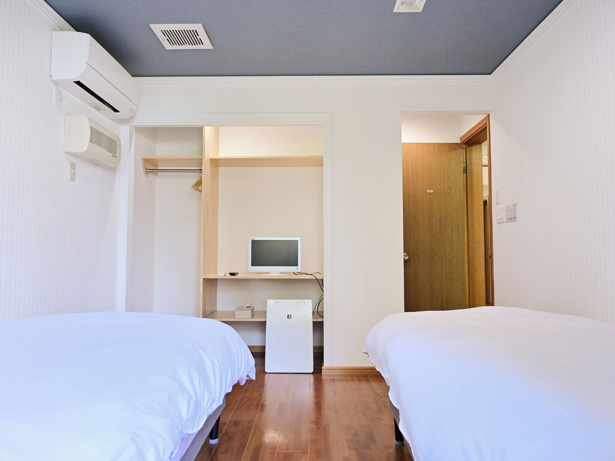 A bedroom with two beds, a desk with a computer, a wardrobe, and a door leading to another room.