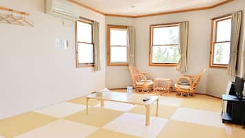 Um quarto com piso xadrez, uma mesa, duas cadeiras de vime, um rack para TV e janelas com cortinas.
