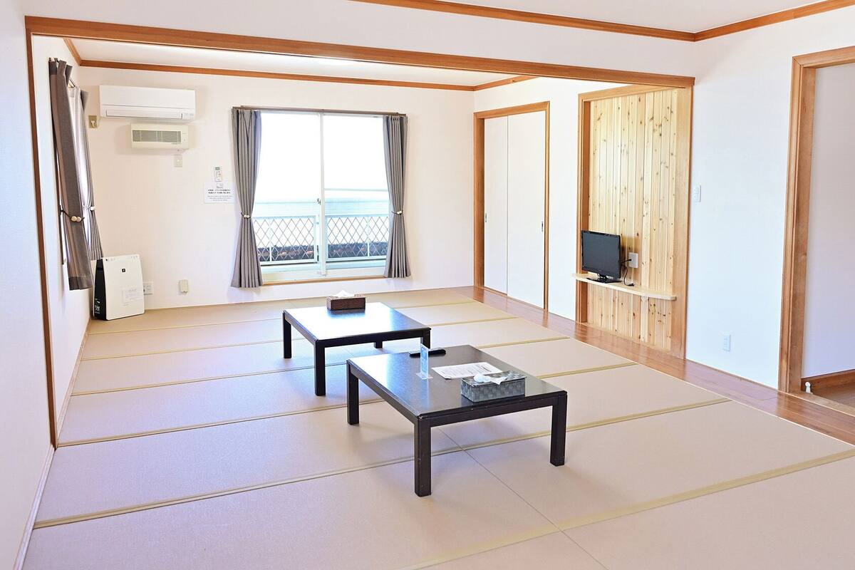 Ein Zimmer mit Tatami-Böden, einer Schiebetür, einem Fernseher auf einer Holzablage und einem fenster mit Vorhängen.