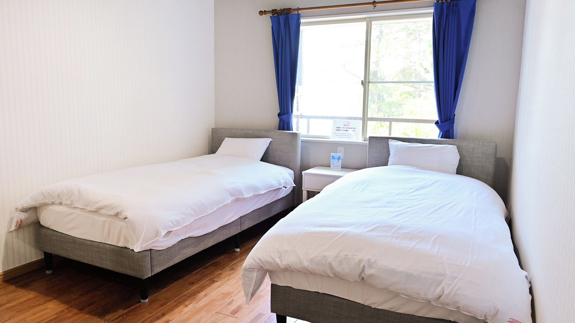 A room with two single beds, a window with curtains, a bedside table, and a nightstand.