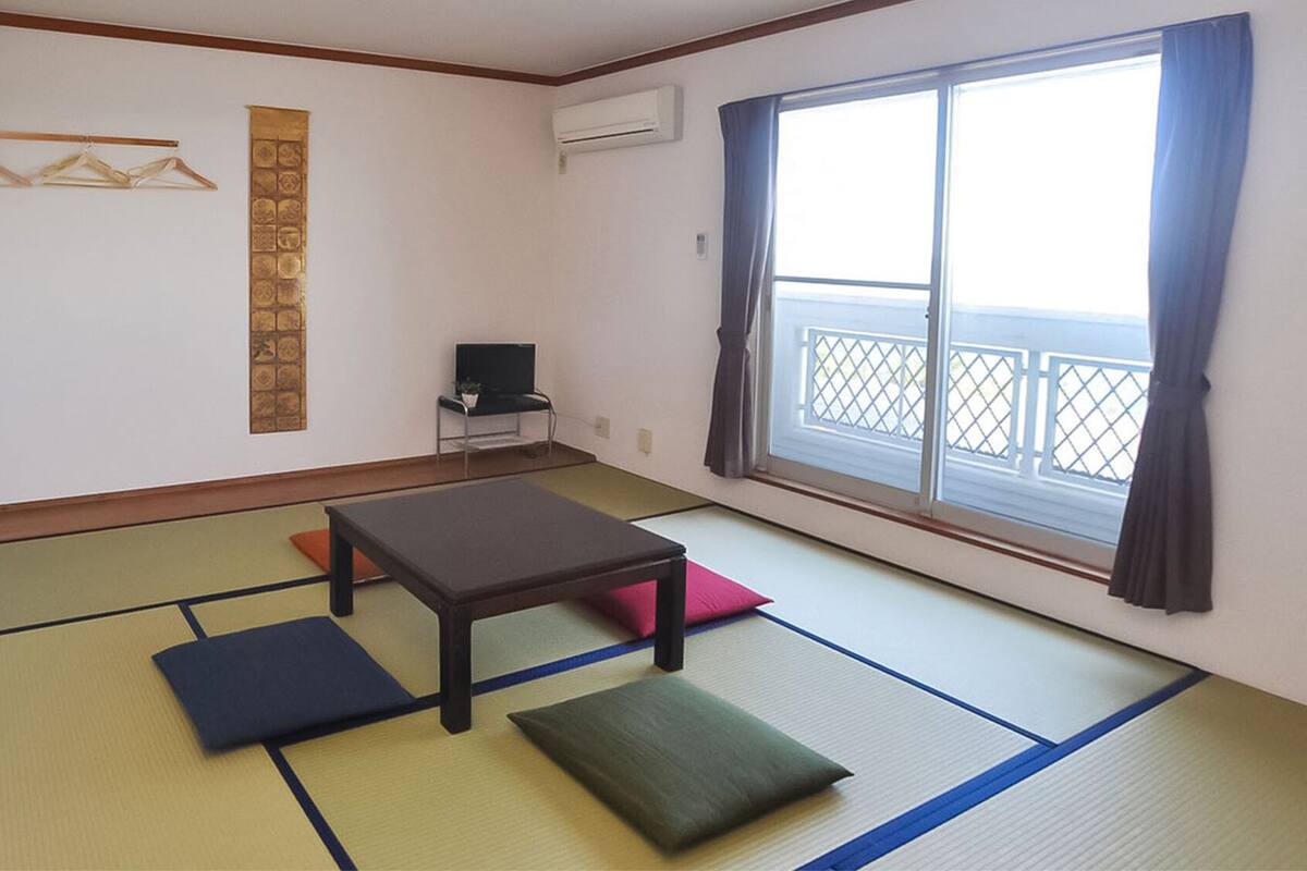 Ein Zimmer mit Tatami-Böden, einem niedrigen Tisch, Kissen, einem Fernseher und einem Fenster mit Blick.