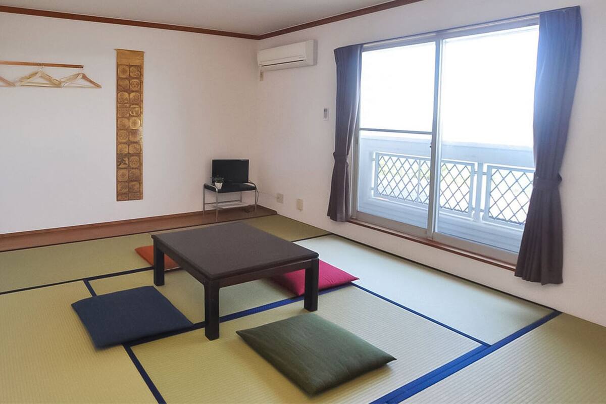 Ein Zimmer mit Tatami-Matten, einem niedrigen Tisch, Kissen, einem Fernseher und einem Fenster mit Blick.