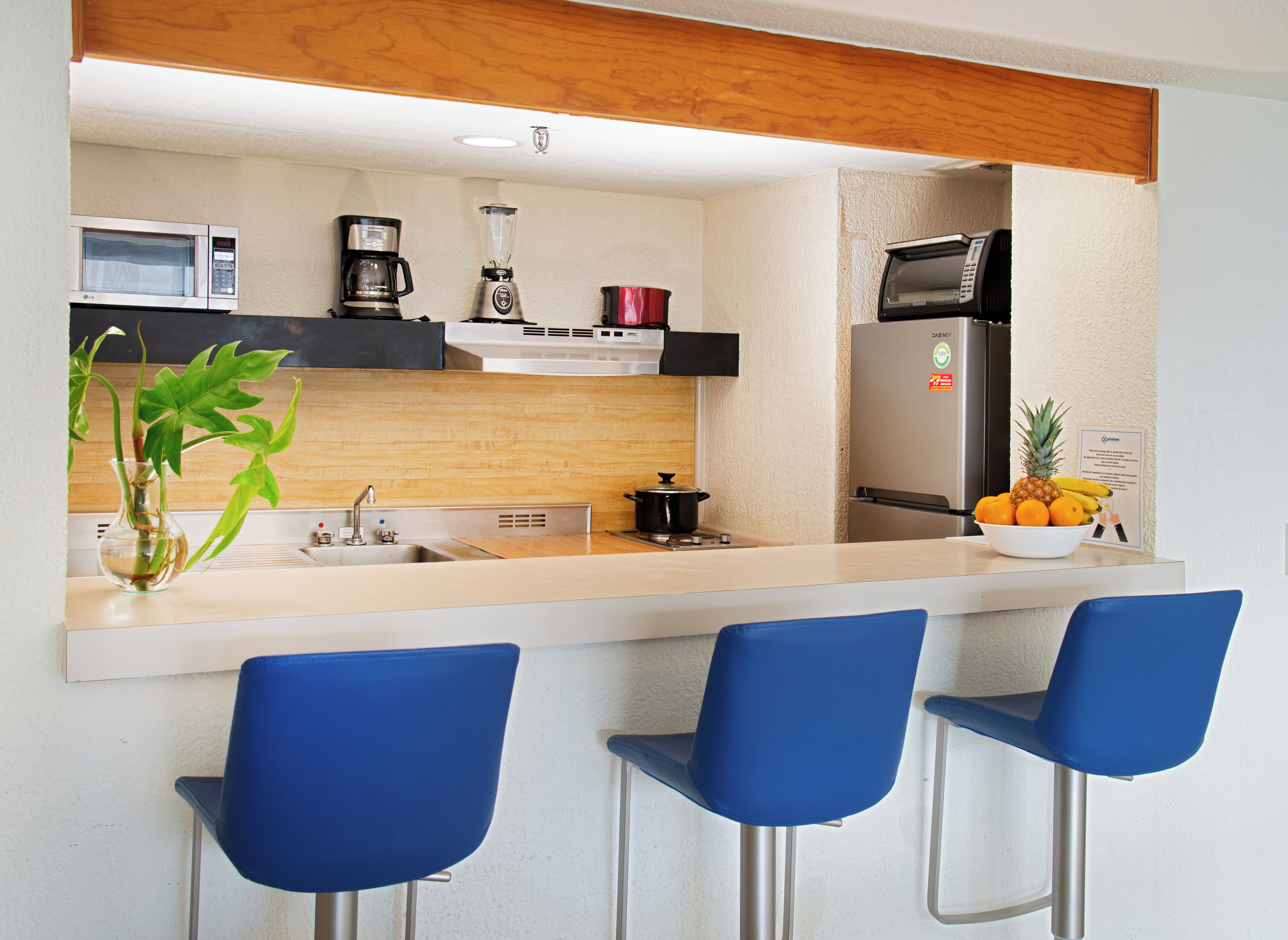 A modern kitchen with a microwave, coffee maker, blender, and a refrigerator. There are three blue bar stools and a bowl of fruit on the counter.