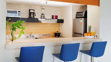 A modern kitchen with a microwave, coffee maker, blender, and a refrigerator. There are three blue bar stools and a bowl of fruit on the counter.