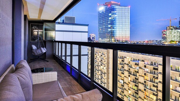 A balcony with a sofa and chair, a view of a cityscape, and a building with lights. - Prestige Skyview Residence – Luxury 2BR • Panoramic Terrace & AC (Warszawa)