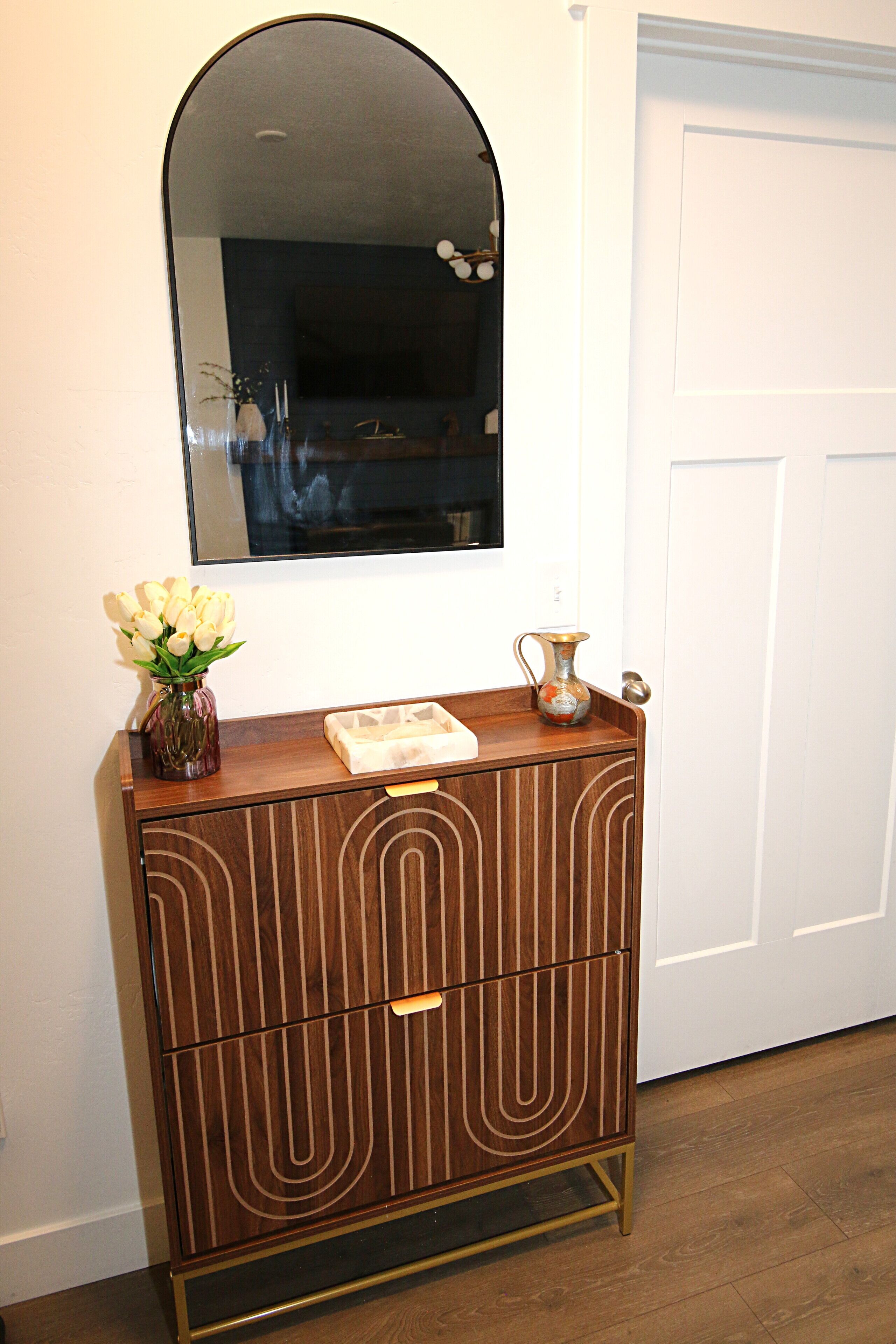 Een houten kast met een spiegel erboven, een vaas met bloemen en een decoratieve kan erop.
