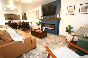 A living room with a sofa, coffee table, fireplace, TV, and dining area. - NEW 6BR Bear Lake Home | 2 Min to Beach | Garage, Pool Access, Game Room | Sleeps 18 (Garden City)