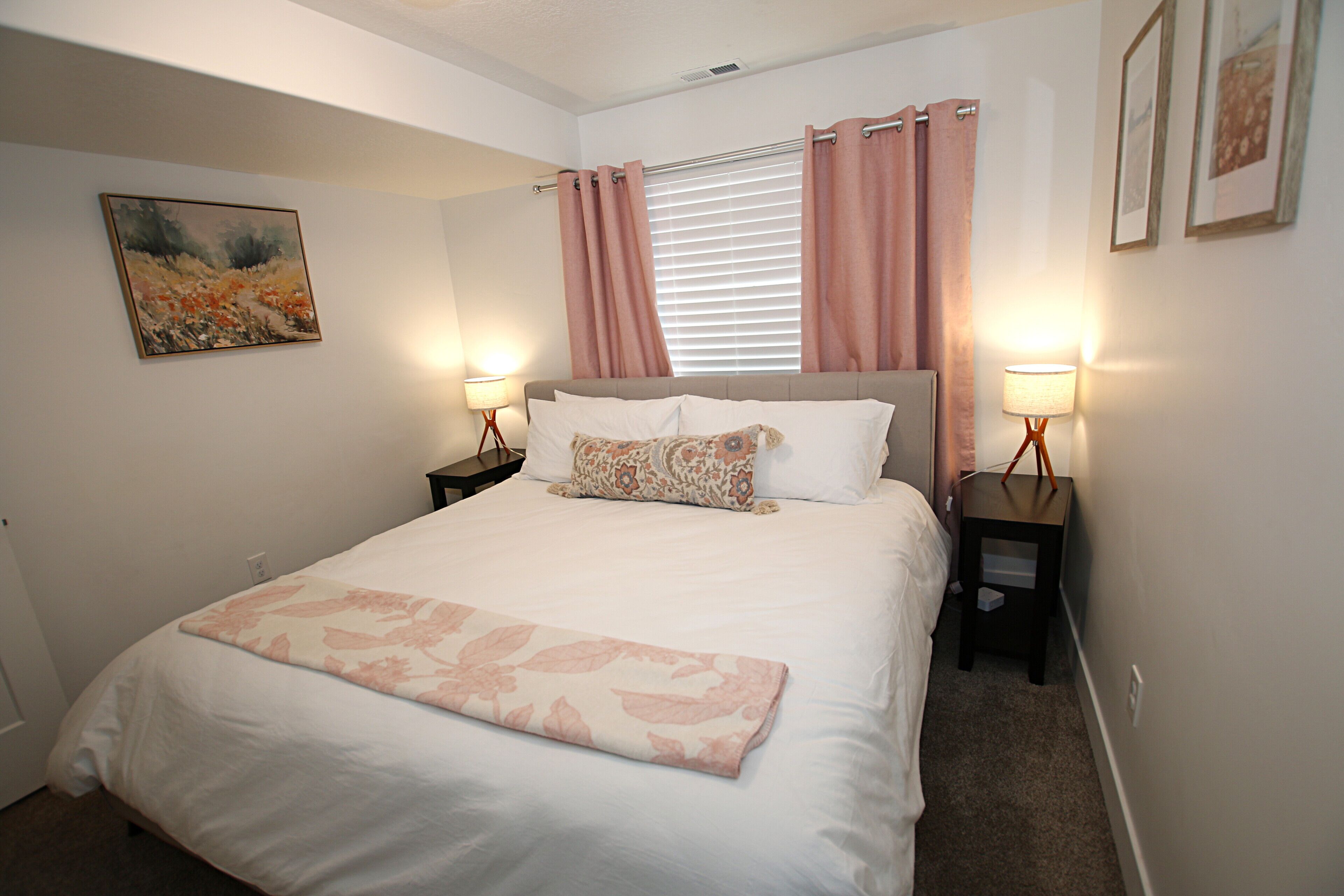 Une chambre à coucher comprenant un lit, deux tables de chevet, une fenêtre avec des rideaux, un tableau accroché au mur et un sol recouvert d’une moquette.