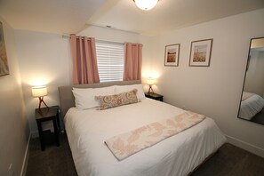 Un dormitorio con cama, dos mesitas de noche, una ventana con cortinas y dos cuadros en la pared.