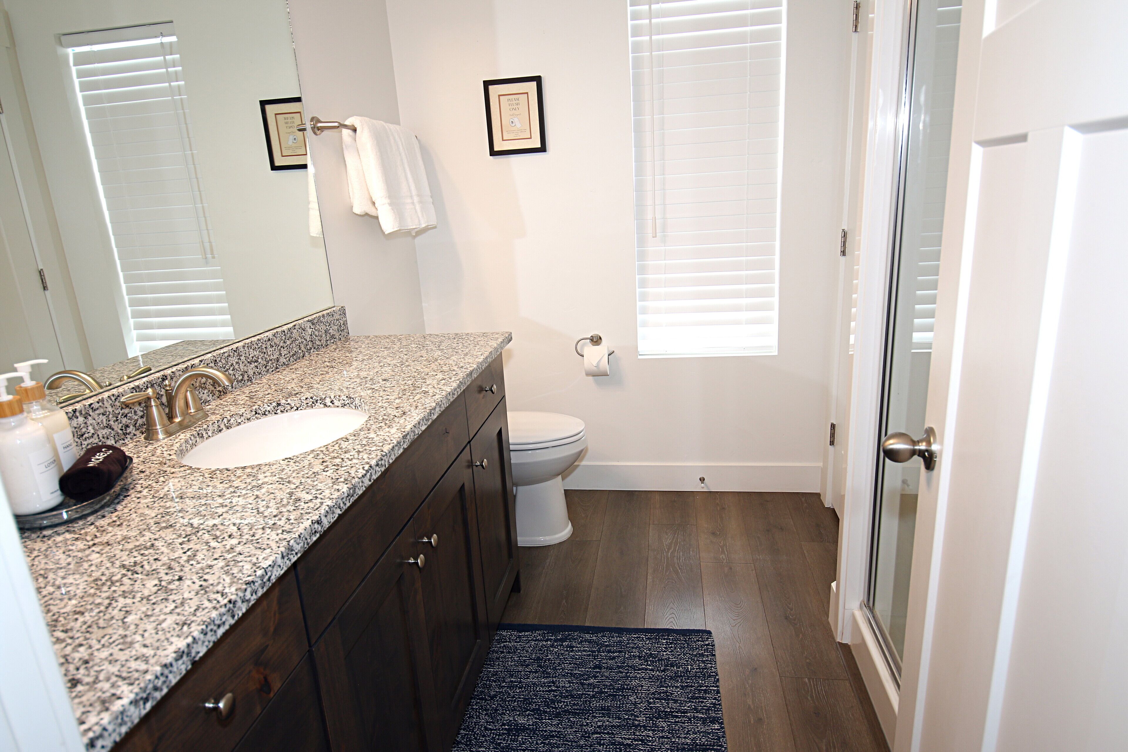 A bathroom with a granite countertop sink, dark wooden cabinets, a white toilet, and a glass shower door.