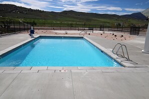 A rectangular swimming pool with a clear blue water, surrounded by a concrete deck and a black metal fence. - NEW 6BR Bear Lake Home | 2 Min to Beach | Garage, Pool Access, Game Room | Sleeps 18 (Garden City)