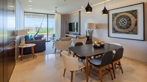 Una habitación de hotel moderna con comedor, un conjunto de sofás, televisor de pantalla plana y un ventanal con vistas al exterior.