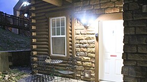 Una cabaña de troncos con fachada de piedra, una puerta con un cartel de 'No Fumar', una ventana y un patio con mesa y sillas.