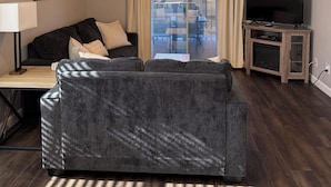 Uma sala de estar com um sofá cinza escuro, uma mesa de centro de madeira, uma TV sobre um móvel de madeira e um ventilador de teto.
