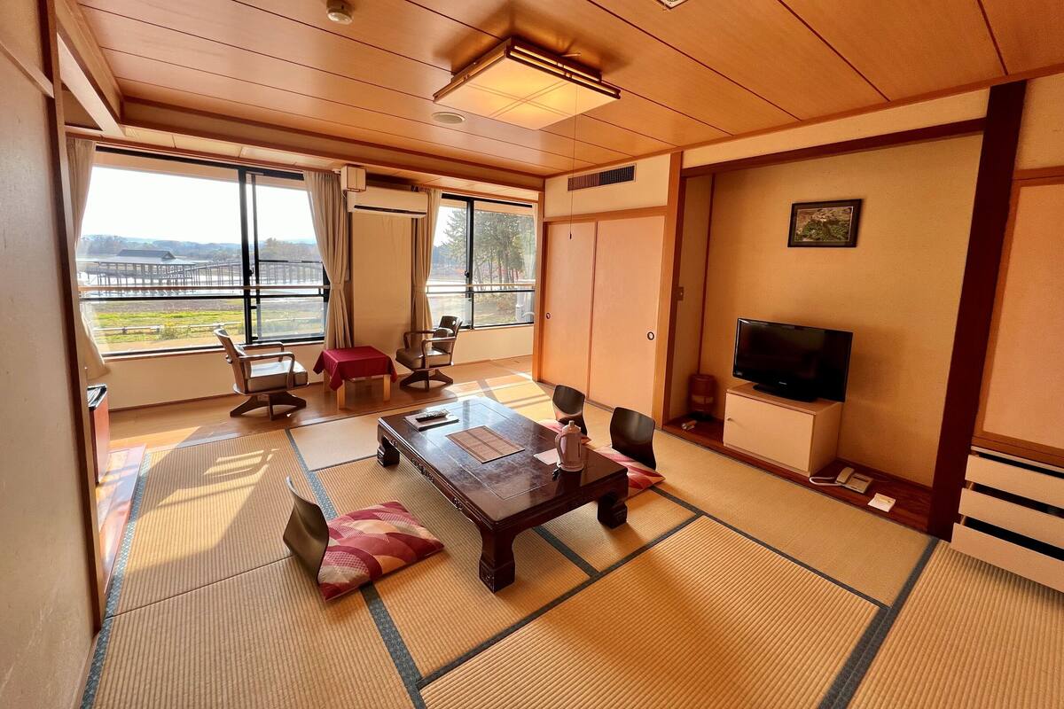A traditional Japanese-style room with tatami flooring, a low table, chairs, a television, and a sliding door leading to a balcony.
