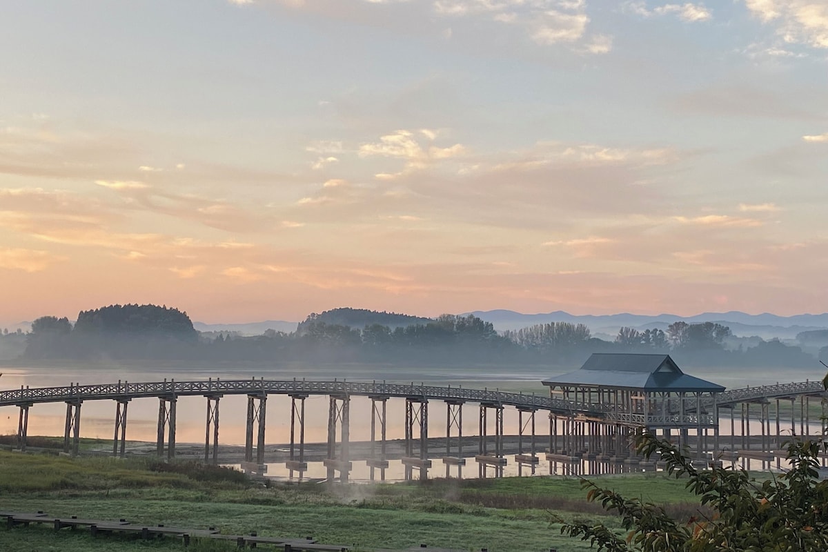 A wooden pier extending over a calm body of water, surrounded by grassy areas and distant hills.