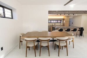 Un espace repas moderne avec une longue table en bois et six chaises, une cuisine en arrière-plan et une grande fenêtre qui laisse entrer la lumière naturelle.