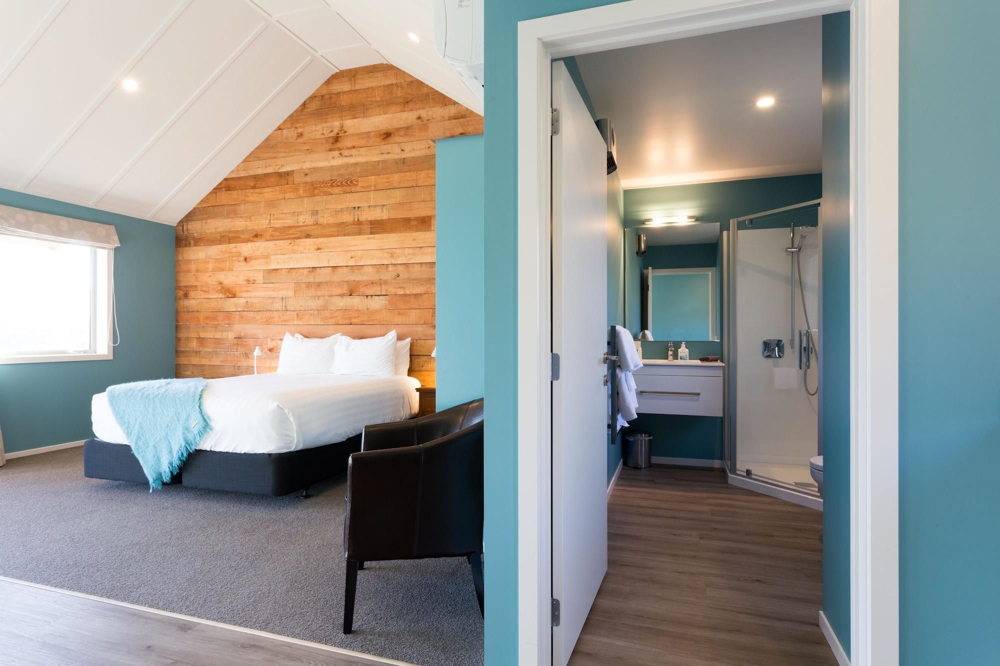 A bedroom with a wooden sloped ceiling, a bed, a chair, and a bathroom with a shower and sink.