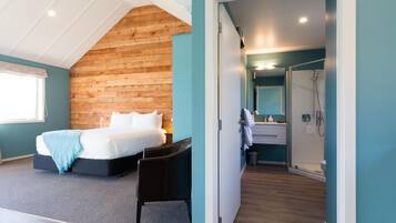 A bedroom with a wooden sloped ceiling, a bed, a chair, and a bathroom with a shower and sink.