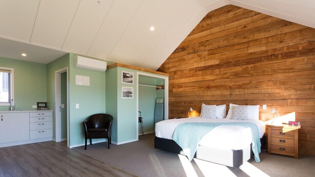 A bedroom with a wooden ceiling, a bed with pillows, a nightstand, and a chair. - Shotover Country Cottages (Lower Shotover)