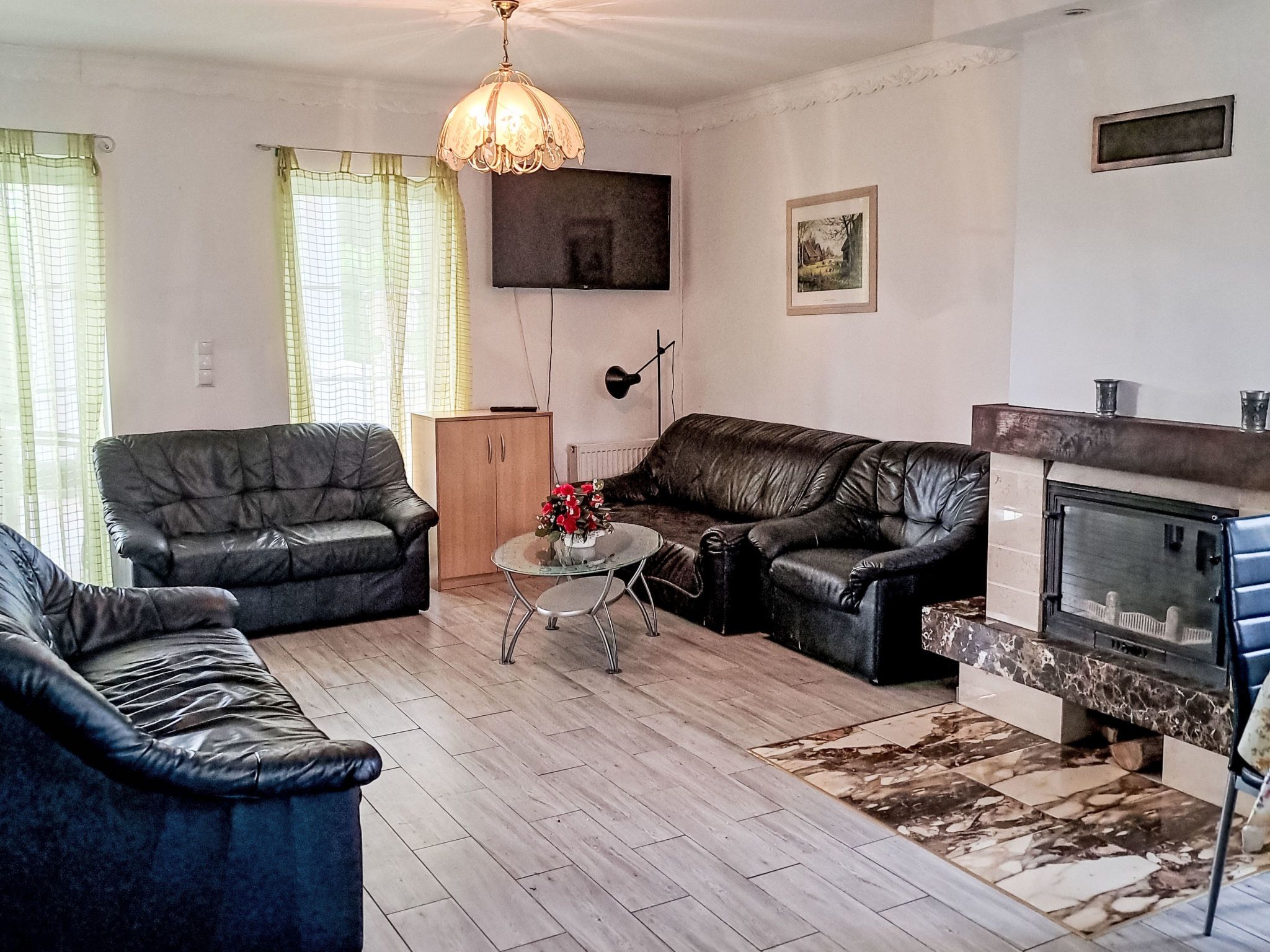 Un salon comprenant deux canapés en cuir noir, une table basse en verre ornée de fleurs rouges, un meuble en bois, un lampadaire noir, une cheminée et un téléviseur fixé au mur.
