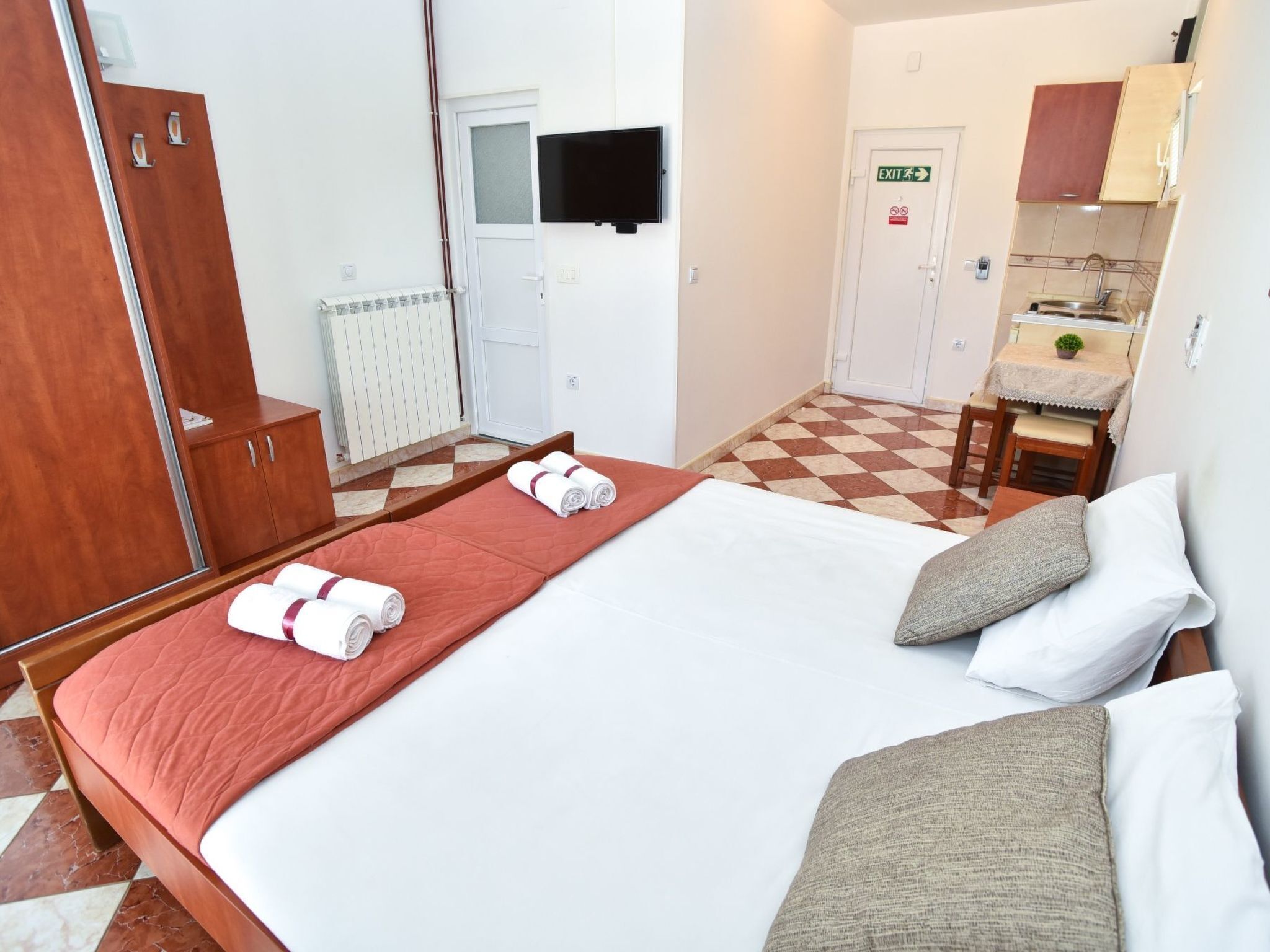 A hotel room with a bed, a wooden wardrobe, a television, a bathroom with a sink and mirror, and a checkered floor.